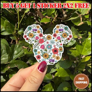 May include: A floral sticker in the shape of a mouse head, featuring a variety of colorful flowers. The sticker is held against a backdrop of green leaves. Text at the top reads "BUY 5 GET 1 STICKER 2X2 FREE". A brown circle with "FAST SHIPPING" is in the bottom right.