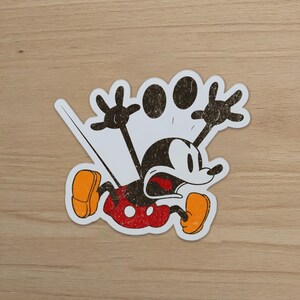 May include: A distressed sticker featuring Mickey Mouse in a distressed, vintage style. Mickey is depicted with a surprised expression, arms raised, and legs bent. The sticker has a white border and is set against a wooden background.