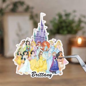 May include: A die-cut sticker featuring a group of illustrated animated princesses arranged in front of a purple castle. The name Brittany is printed in black script at the bottom. The sticker is held by silver metal tweezers.