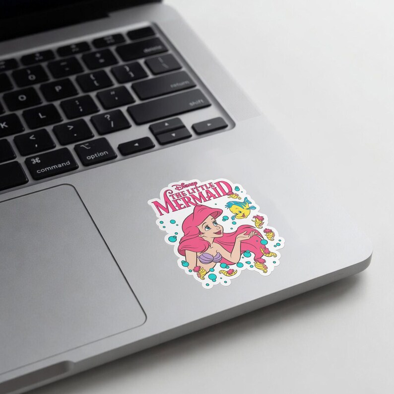 Op de afbeelding: Een gestanste sticker met Ariel uit De Kleine Zeemeermin met lang rood haar, een paarse schelpentop en een groene staart. Het ontwerp bevat Botje, kleine zeepaardjes en blauwe bubbels, met de tekst Disney The Little Mermaid in het roze.