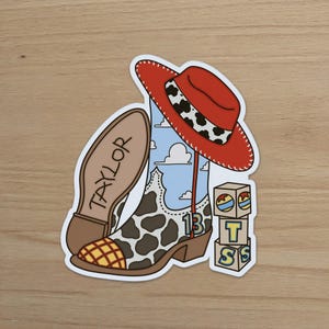 Puede incluir: Una pegatina que muestra una bota de vaquero con estampado de manchas de vaca, un fondo de cielo azul con nubes y un sombrero de vaquero rojo. La suela dice TAYLOR, el n&uacute;mero 13 est&aacute; en el lateral y hay una pila de tres cubos con las letras T y S.