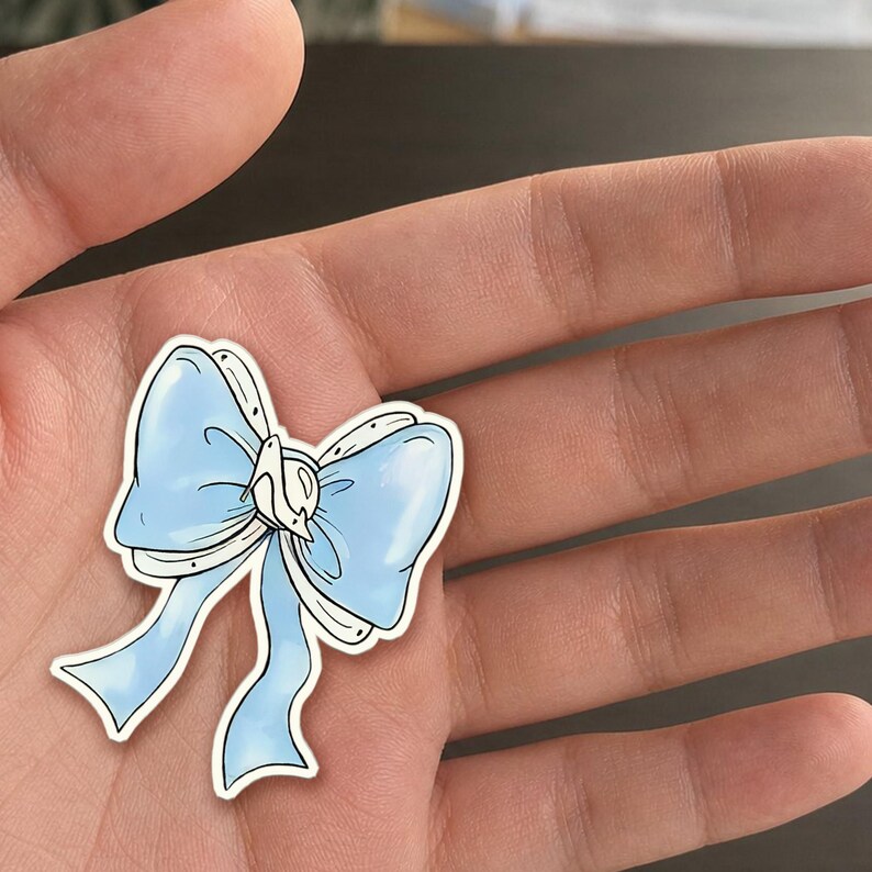 May include: A light blue bow sticker with white accents and a white center. The sticker is held in a hand, showcasing the delicate design and color palette. The bow has a classic shape with flowing ribbons.