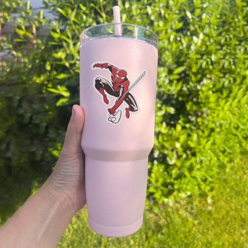 May include: A pale pink tumbler with a clear lid and a matching pink straw. A sticker of Spider-Man in a dynamic pose is affixed to the tumbler. The tumbler is designed to hold a large quantity of liquid.