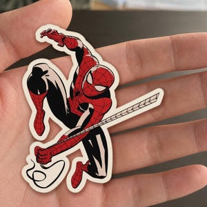 May include: A sticker featuring a dynamic illustration of Spider-Man in a classic pose. The character is depicted in his iconic red and black suit, with a web extending from his hand. The sticker has a white border.