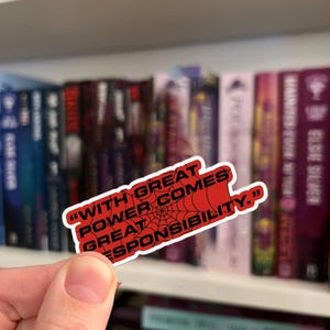 May include: A red sticker with the quote "WITH GREAT POWER COMES GREAT RESPONSIBILITY" in white text, featuring a spiderweb design. The sticker is held in front of a bookshelf filled with books.