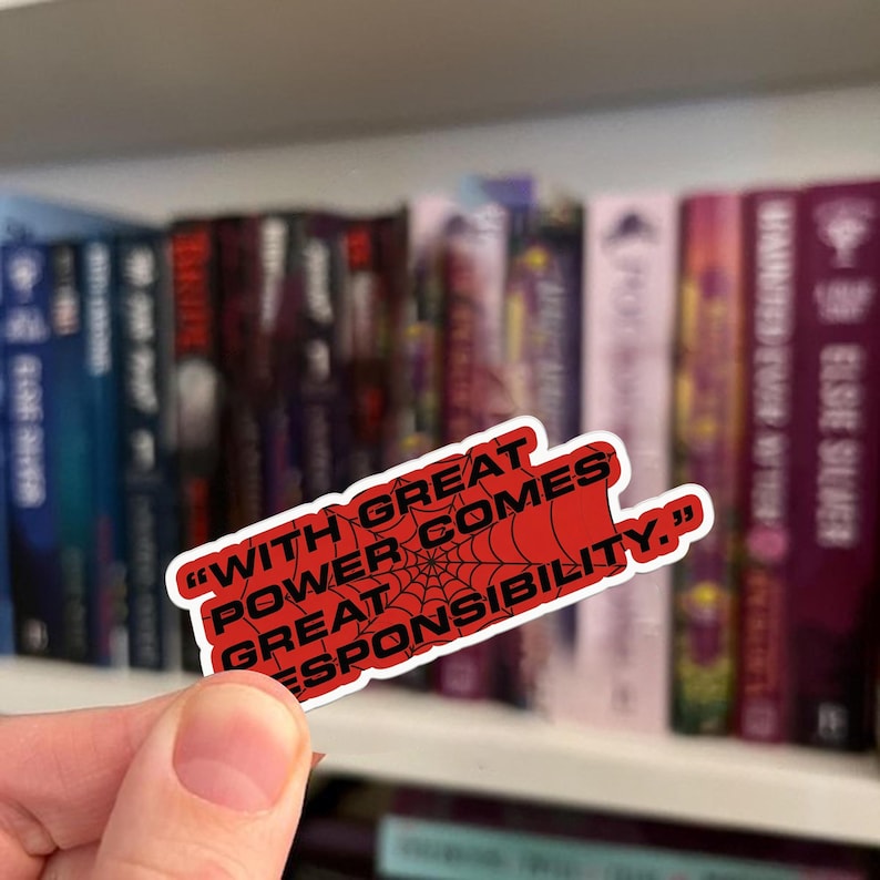 May include: A red sticker with the quote "WITH GREAT POWER COMES GREAT RESPONSIBILITY" in white text, featuring a spiderweb design. The sticker is held in front of a bookshelf filled with books.