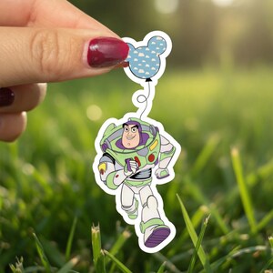 May include: A sticker featuring Buzz Lightyear from Toy Story, running with a Mickey Mouse-shaped balloon. Buzz is in his green, white, and purple spacesuit. The balloon is light blue with white cloud shapes. The sticker is held against a blurred green background.