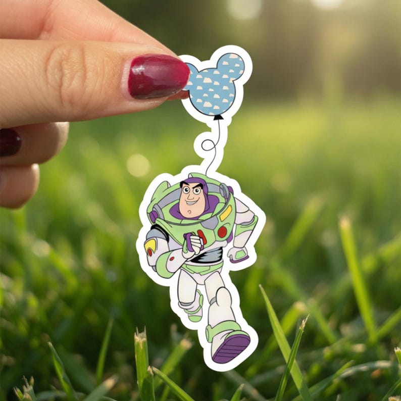 May include: A sticker featuring Buzz Lightyear from Toy Story, running with a Mickey Mouse-shaped balloon. Buzz is in his green, white, and purple spacesuit. The balloon is light blue with white cloud shapes. The sticker is held against a blurred green background.