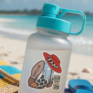 Puede incluir: Una botella de agua esmerilada con tapa turquesa y una pegatina que muestra una bota de vaquero con estampado de vaca, un sombrero rojo con banda de estampado de vaca, el nombre Taylor, el n&uacute;mero 13 y tres bloques con las letras T, S y S.