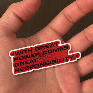 May include: A red sticker with white trim featuring the quote "WITH GREAT POWER COMES GREAT RESPONSIBILITY." The text is in black, with a spiderweb design in the center. The sticker is held in a hand.