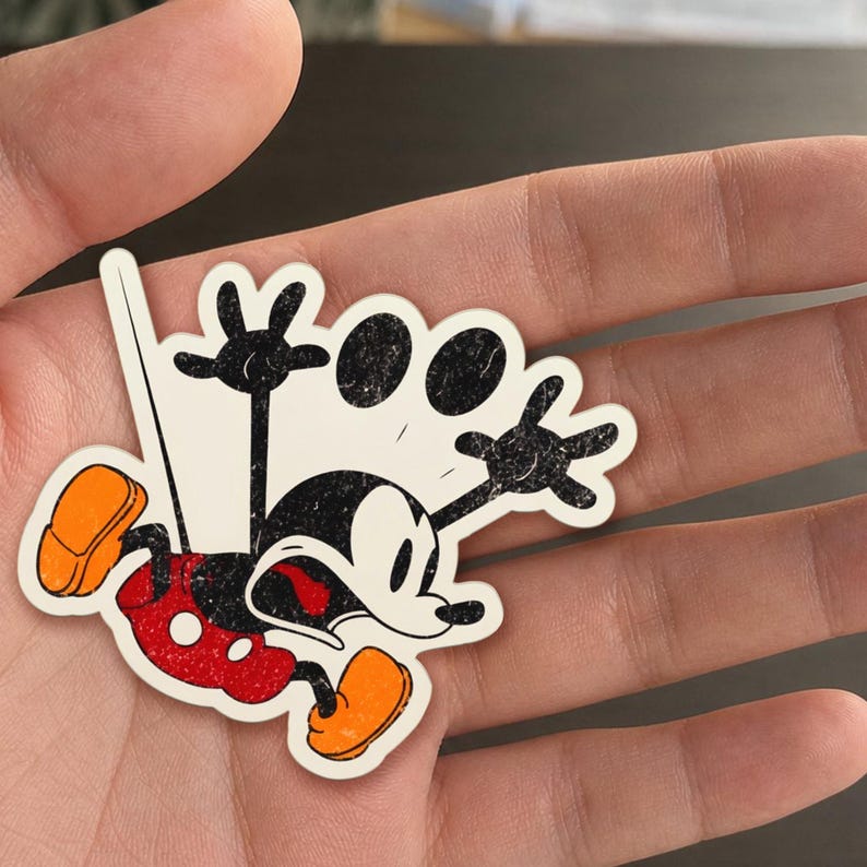 May include: A distressed sticker featuring Mickey Mouse in a falling pose. The sticker has a white background with black, red, and orange accents. Mickey's iconic ears are visible, along with his red shorts and orange shoes.
