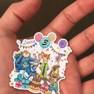 May include: A die-cut sticker featuring Zootopia characters including Judy Hopps, Nick Wilde, Chief Bogo, and Gazelle. The design includes a birthday cake, colourful balloons, confetti, and the text Lucas and the number 3 in a balloon.