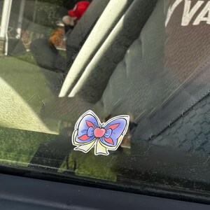 May include: A sticker of a cartoon bow with a heart in the center. The bow has blue, red, and yellow accents. The sticker is adhered to a car window.