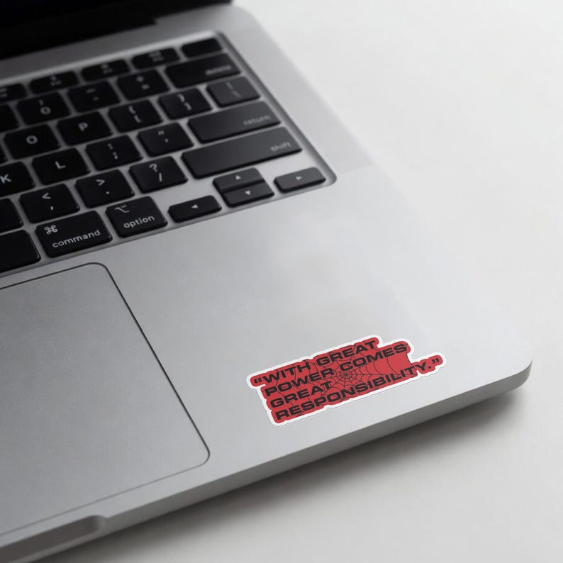 May include: A red sticker with the quote "WITH GREAT POWER COMES GREAT RESPONSIBILITY." The sticker is rectangular with a white border and is placed on a silver laptop.