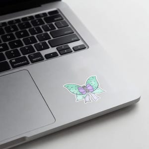 May include: A sticker featuring a whimsical butterfly design. The butterfly has light green wings with purple accents and a purple bow. The sticker is placed on a silver laptop.