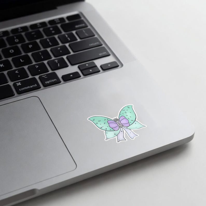 May include: A sticker featuring a whimsical butterfly design. The butterfly has light green wings with purple accents and a purple bow. The sticker is placed on a silver laptop.