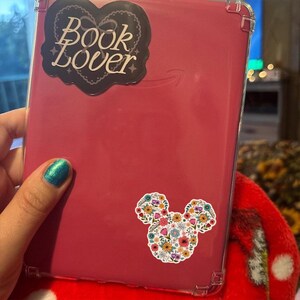 May include: A red tablet with a clear protective case. The tablet has two stickers: one with the words "Book Lover" and a heart design, and another in the shape of a mouse head filled with colorful flowers.