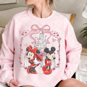 May include: Pink sweatshirt featuring a graphic of Mickey and Minnie Mouse in front of a castle. Minnie holds a heart-shaped box. The design includes a pink bow, flowers, and a decorative border. The characters are wearing red and black outfits.