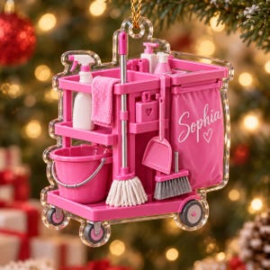 May include: A clear acrylic Christmas ornament shaped like a pink cleaning cart. The cart features a bucket, mop, broom, dustpan, spray bottle, and a cloth. The side of the cart is printed with the name Sophia and a small heart icon.