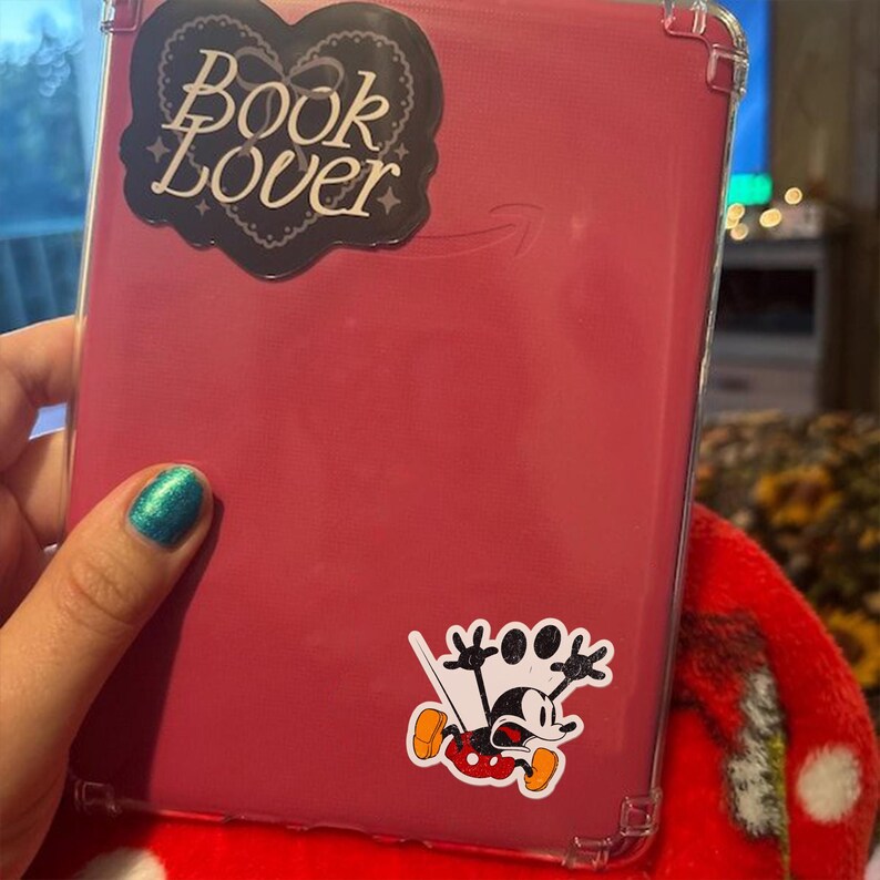 May include: A pink e-reader case with a clear protective cover. The case has a "Book Lover" sticker in a heart shape and a Mickey Mouse sticker. The e-reader is held by a person with teal nail polish.