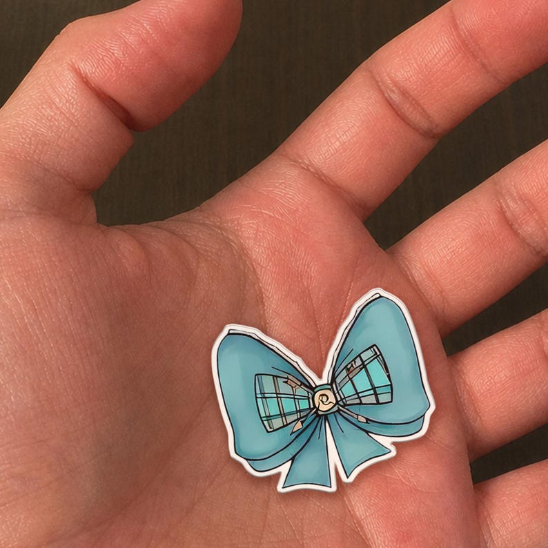 May include: A light blue bow-shaped sticker with a white border. The bow has a plaid pattern in the center and a small gold-colored embellishment. The sticker is held in a person's hand.
