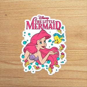 Op de afbeelding: Een gestanste sticker met Ariel uit Disney's De Kleine Zeemeermin met felroze haar, een paarse schelpentop en de gele vis Botje. Het ontwerp bevat verschillende kleine gele zeepaardjes en turquoise bubbels, met de tekst Disney THE LITTLE MERMAID in het roze bovenaan gedrukt.