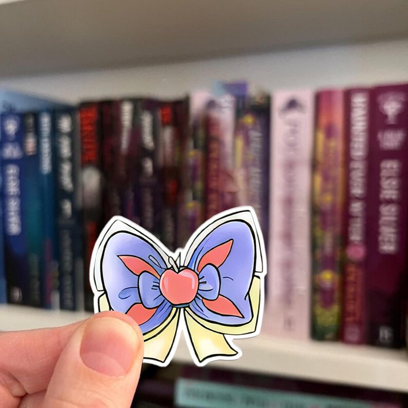 May include: A sticker featuring a cartoon bow with a red apple center. The bow has light blue, yellow, and coral accents. The sticker is held in front of a bookshelf filled with books.