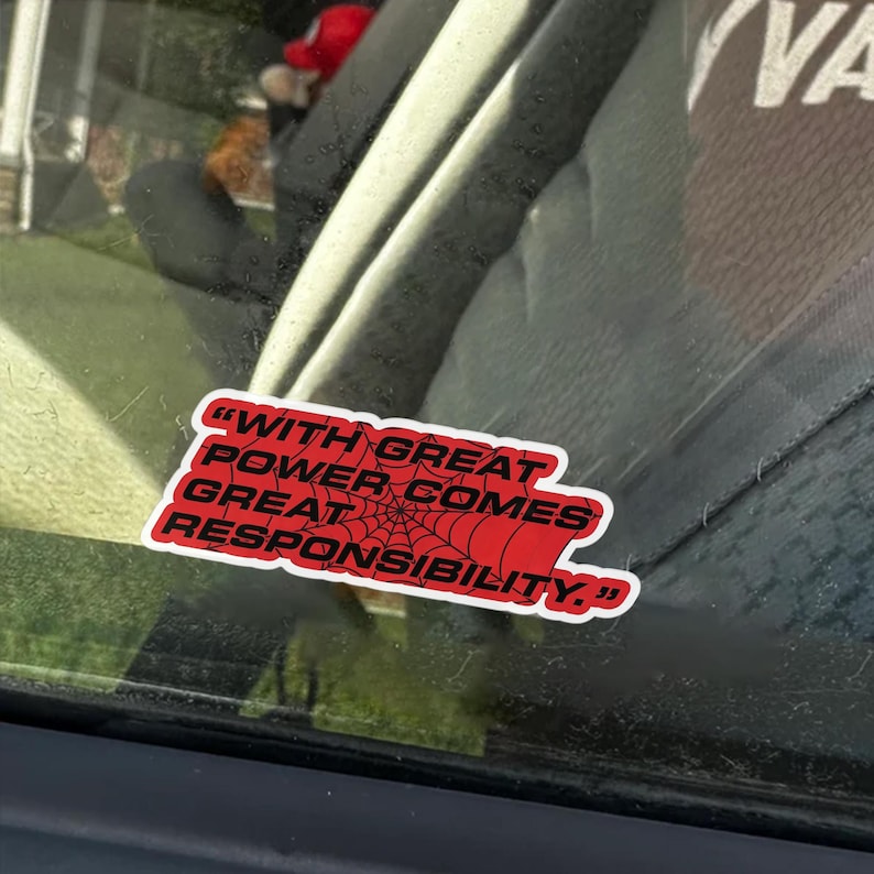 May include: A red sticker with the quote "WITH GREAT POWER COMES GREAT RESPONSIBILITY" in white text. The sticker has a spiderweb design and is affixed to a car window.