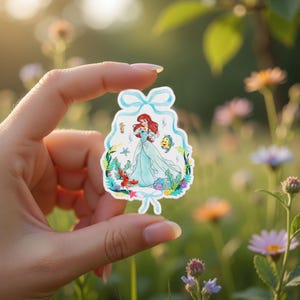 Peut inclure: Un autocollant d&eacute;coup&eacute; repr&eacute;sentant une illustration &agrave; l'aquarelle d'une princesse en robe turquoise fluide, avec des cheveux roux et un poisson jaune. L'autocollant est entour&eacute; de plantes et de cr&eacute;atures aquatiques, avec un n&oelig;ud bleu clair en haut.