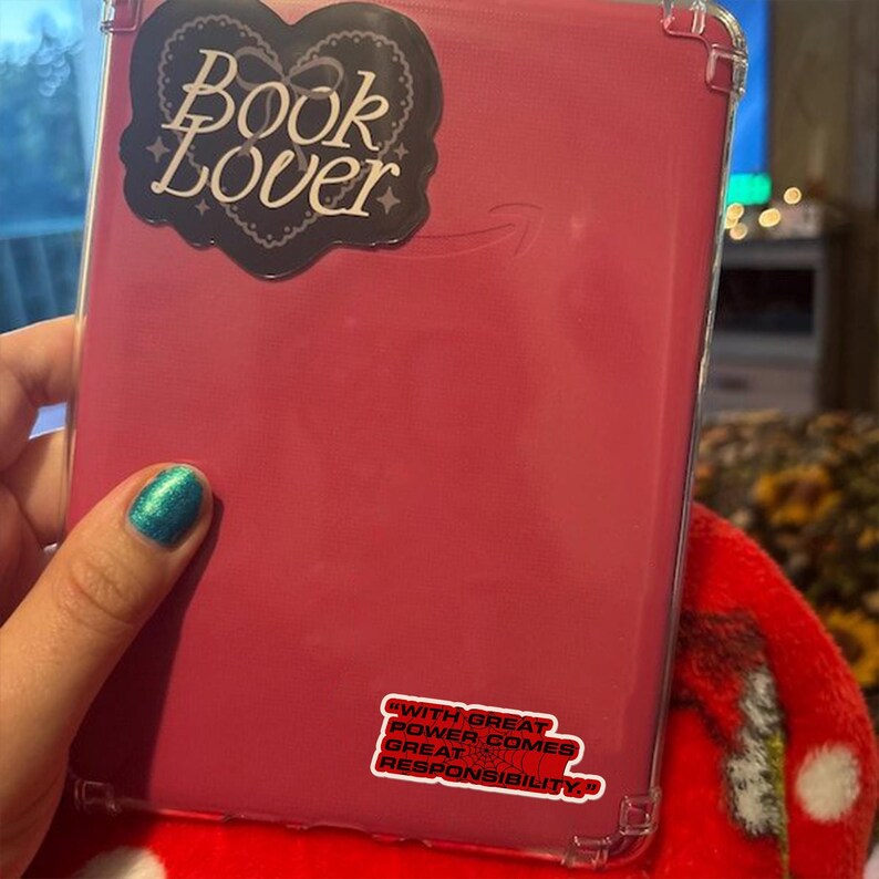 May include: A pink e-reader case with a clear protective cover. The case has a sticker that says "Book Lover" in a heart-shaped design. A smaller sticker reads "With great power comes great responsibility."