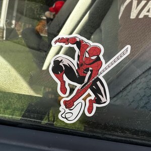 May include: A sticker of Spider-Man in a dynamic pose, with a red and black costume. The sticker has a white border and depicts the superhero with his web-shooters and web.