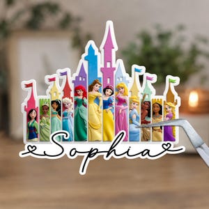 May include: A decorative cutout featuring a colorful castle silhouette with various animated princesses standing in front of individual towers. The name Sophia is written in black cursive script below the castle, flanked by two small heart outlines.