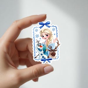 以下が含まれることがあります： アナと雪の女王のエルサ、雪だるまのオラフ、トナカイのスヴェンが描かれた長方形のステッカー。キャラクターは、青と白の雪の結晶をテーマにしたボーダーに、青いリボンが添えられています。エルサは手を振っており、オラフは人参の鼻をしています。