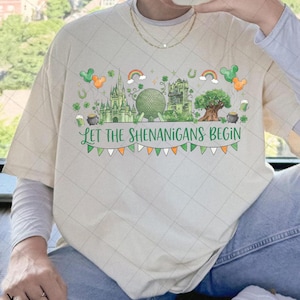 Puede incluir: Camiseta color crema con un diseño verde y naranja que presenta un castillo, una gran esfera verde, un árbol y el texto "LET THE SHENANIGANS BEGIN". El diseño incluye arcos iris, tréboles y globos con forma de Mickey Mouse.