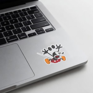 May include: A distressed sticker featuring Mickey Mouse in a running pose. The cartoon character is depicted in black, red, and orange, with a white background. The sticker is placed on a silver laptop.