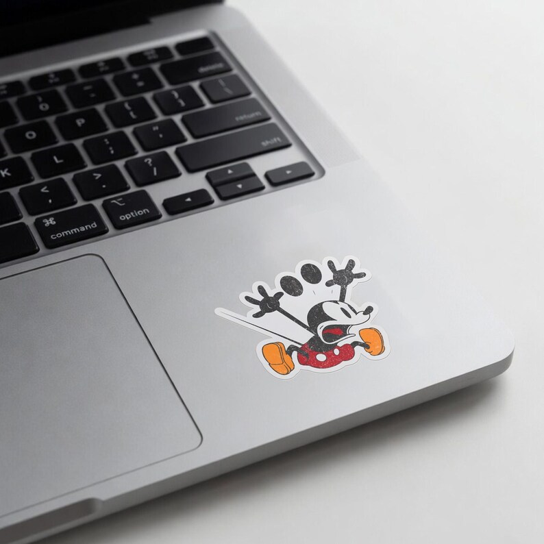 May include: A distressed sticker featuring Mickey Mouse in a running pose. The cartoon character is depicted in black, red, and orange, with a white background. The sticker is placed on a silver laptop.