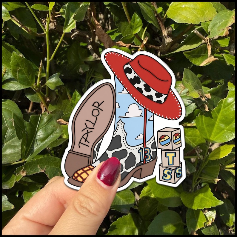 Puede incluir: Una pegatina troquelada que muestra una bota de vaquero con un patr&oacute;n de cielo azul y nubes, una superposici&oacute;n de estampado de vaca y el n&uacute;mero 13. Un sombrero de vaquero rojo con una banda de estampado de vaca descansa sobre la bota. Una suela de bota marr&oacute;n dice TAYLOR. Tres bloques apilados muestran las letras T, S, S.