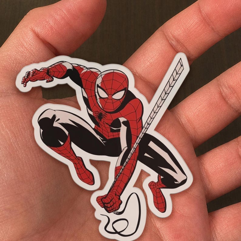 May include: A sticker featuring a comic book style illustration of Spider-Man in a dynamic pose. The character is depicted in his classic red and black suit, with a web-slinging accessory. The sticker is on a white background.