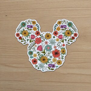 May include: A die-cut sticker in the shape of Mickey Mouse's head, filled with a colorful floral design. The flowers include red, pink, yellow, and blue blooms, with green leaves and stems. The sticker is on a wooden surface.