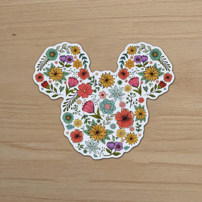 May include: A die-cut sticker in the shape of Mickey Mouse's head, filled with a colorful floral design. The flowers include red, pink, yellow, and blue blooms, with green leaves and stems. The sticker is on a wooden surface.