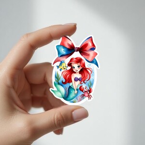 Peut inclure: Un autocollant repr&eacute;sentant une illustration &agrave; l'aquarelle d'une sir&egrave;ne aux cheveux roux, avec un soutien-gorge en coquillage violet et une queue turquoise. Un n&oelig;ud rouge et bleu est en haut. Un poisson jaune et un crabe rouge sont &eacute;galement dans l'image.