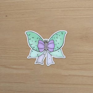 May include: A sticker featuring a whimsical butterfly bow design. The bow has light green wings with a textured pattern, a purple center, and white and light purple ribbons. The sticker is on a wooden surface.