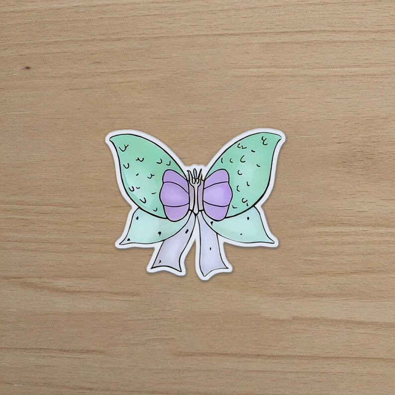 May include: A sticker featuring a whimsical butterfly bow design. The bow has light green wings with a textured pattern, a purple center, and white and light purple ribbons. The sticker is on a wooden surface.
