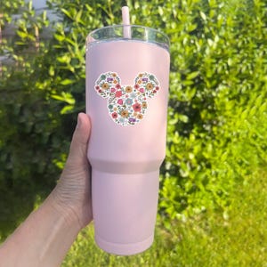 May include: A light pink tumbler with a clear lid and a pink straw. The tumbler features a floral Mickey Mouse head design in various colors. The tumbler is held by a hand against a backdrop of green foliage.