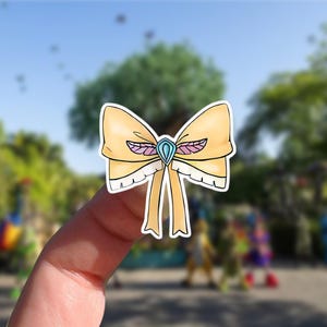 May include: A yellow bow sticker with a blue gem in the center and pink and white accents. The bow has a ribbon design and is held up by a finger.