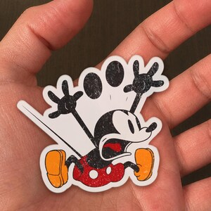 May include: A distressed sticker featuring a cartoon mouse character in a state of panic. The mouse has black and white features, red shorts, and orange shoes. The sticker is white with black outlines and a distressed texture.