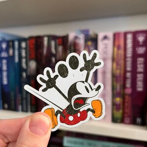 May include: A distressed-style sticker featuring Mickey Mouse in a running pose with a look of surprise. The sticker has a white background with black and red accents, including Mickey's iconic red shorts and yellow shoes. The word "look" is above Mickey's head.