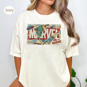 May include: Ivory-colored t-shirt featuring a comic book-style graphic with the word "MARVEL" in red and white. The graphic includes images of superheroes and action words like "SMASH!" and "KAPOW!"