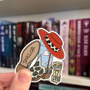 Puede incluir: Una pegatina troquelada que muestra una bota de vaquero con estampado de vaca, un cielo azul con nubes blancas y el n&uacute;mero 13. Un sombrero de vaquero rojo con una banda de estampado de vaca descansa encima. La suela dice TAYLOR y hay tres cubos.