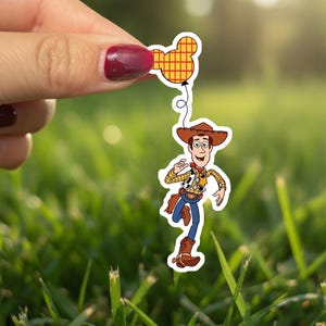 May include: A sticker of the character Woody from Toy Story, wearing a cowboy hat, vest, and blue jeans, running and holding a Mickey Mouse balloon. The balloon is yellow and red. The background is green grass.